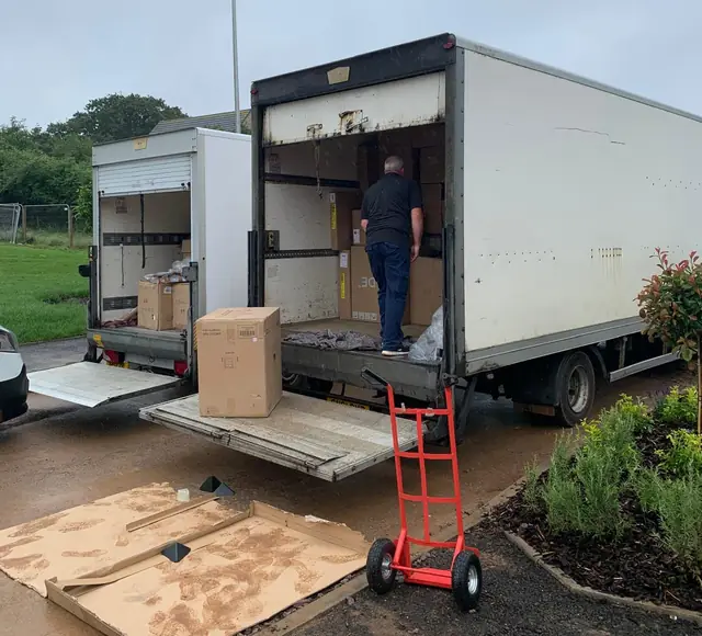 Two Luton vans with tail lifts lowered, a large box on one lift and a red sack truck on the pavement while a team member loads the van