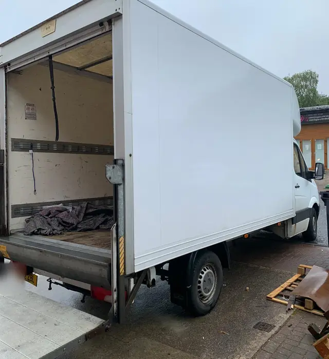 White Luton van seen from the side and rear, with the rear shutter up and the tail lift lowered to the ground